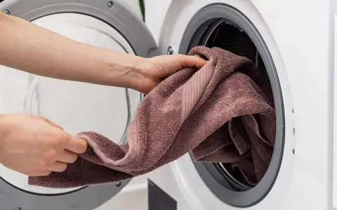 Manos colocando una toalla marrón en una lavadora. Aprende como lavar toallas para que queden suaves y esponjosas.
