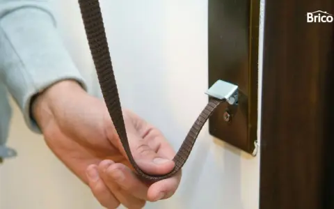 Mano sujetando una cinta marrón que se ha roto la cuerda de la persiana, enganchada a un soporte metálico.