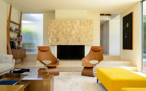 Dos sillas de madera con cojines de cuero marrón, un sofá amarillo y una chimenea de piedra, todo con un inconfundible estilo mid century.