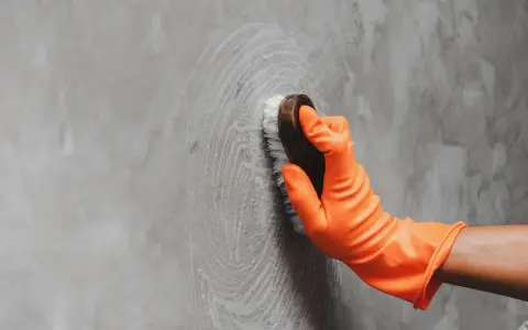 Mano con guante naranja limpiando una pared de concreto con un cepillo, dejando círculos de espuma.