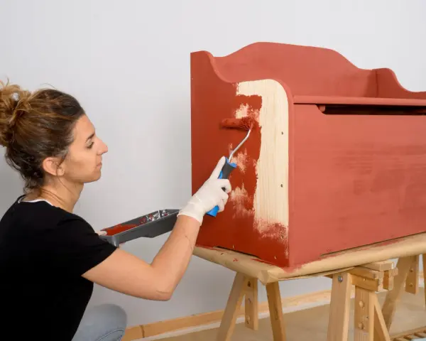 Mujer con guantes blancos pinta un mueble de madera con rodillo, cubriendo el barniz con pintura roja.