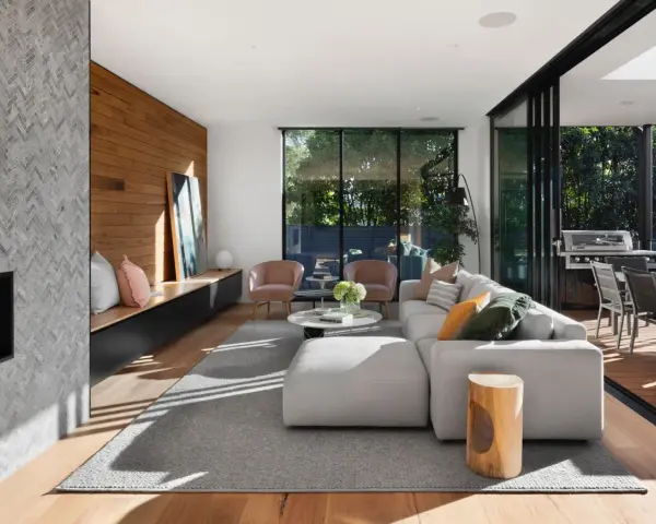 Interior de casa de madera con chimenea de mosaico, sofá modular gris, sillones rosas y acceso a terraza con barbacoa.