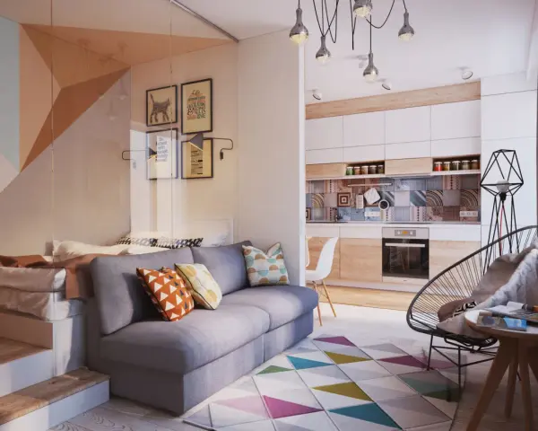 Un sofá gris con cojines coloridos. Detrás, una cama elevada y una pared con cuadros. La cocina moderna y una silla de diseño completan el espacio.