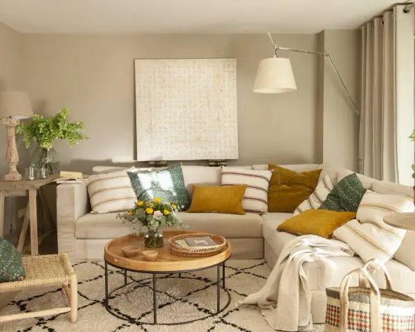 Sofá beige con cojines variados, mesa de centro redonda de madera, alfombra estampada y una silla de mimbre. El color topo cual es, se ve en las paredes y cortinas.
