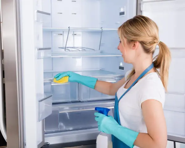 Mujer con guantes azules limpia el interior de una nevera vacía con una esponja amarilla y spray.