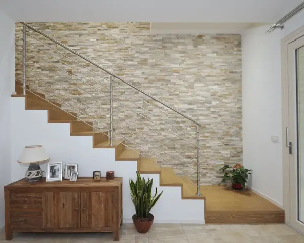 Elegante escalera con barandilla metálica y una pared de piedra natural para decorar pared subida escalera. Un mueble de madera y plantas completan la escena.