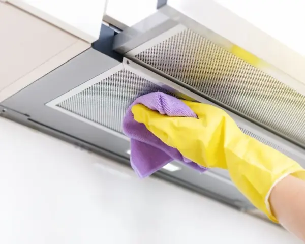 Mano con guante amarillo y paño morado limpiando el interior de la campana extractora, eliminando grasa y suciedad para un hogar reluciente.