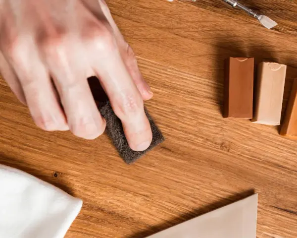 Mano lijando un parquet rayado con una esponja. Cerca, barras de cera para madera y un paño blanco.