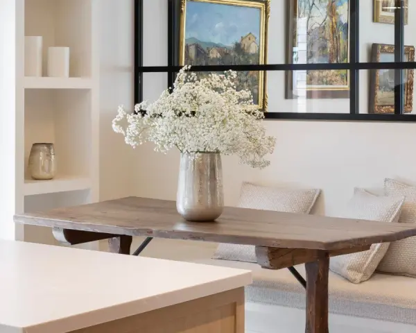 Un jarrón con flores blancas decora la mesa de madera rústica. El espacio alrededor de la mesa comedor es acogedor, con un banco tapizado y cojines.