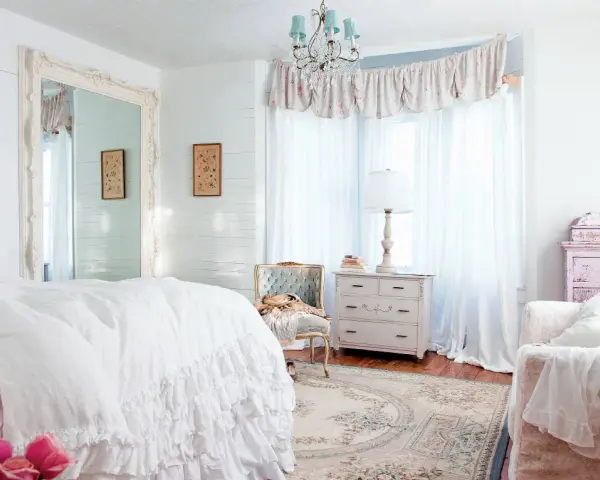 Dormitorio con cama blanca con volantes, espejo grande, cómoda antigua y un sillón. Estilo shabby chic.