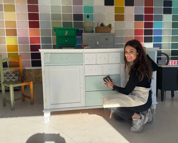 Mujer sonríe mientras lija un mueble para pintar. Detrás, una pared con muestras de colores.