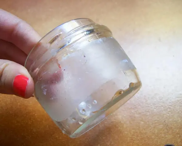 Mano con uña roja sostiene frasco de vidrio con residuos de pegamento. Aprende como quitar el pegamento de una pegatina.