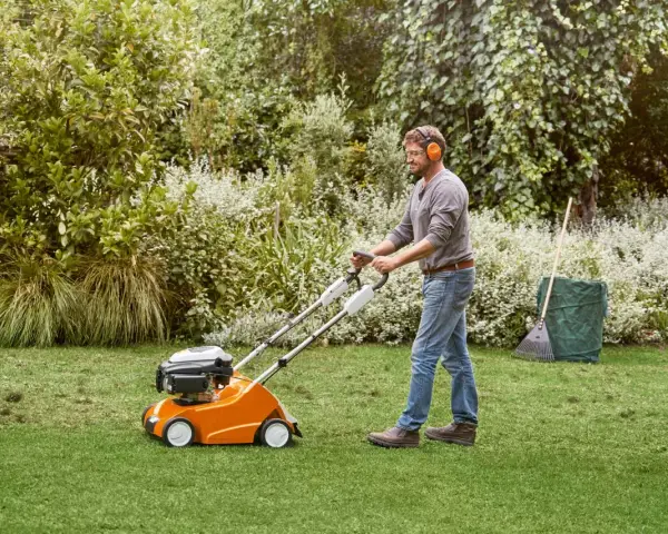 Hombre con auriculares usa un escarificador naranja para airear el césped en un jardín verde.