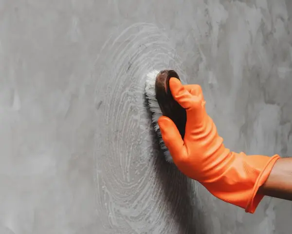 Mano con guante naranja limpiando una pared de concreto con un cepillo, dejando círculos de espuma.