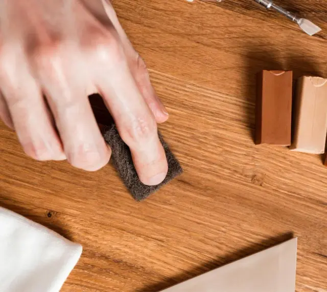 Mano lijando un parquet rayado con una esponja. Cerca, barras de cera para madera y un paño blanco.