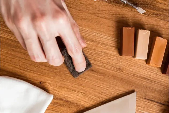 Mano lijando un parquet rayado con una esponja. Cerca, barras de cera para madera y un paño blanco.