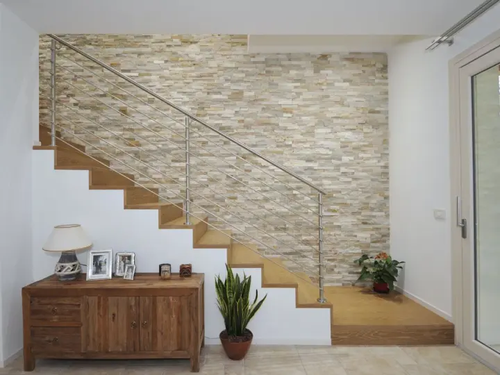 Elegante escalera con barandilla metálica y una pared de piedra natural para decorar pared subida escalera. Un mueble de madera y plantas completan la escena.