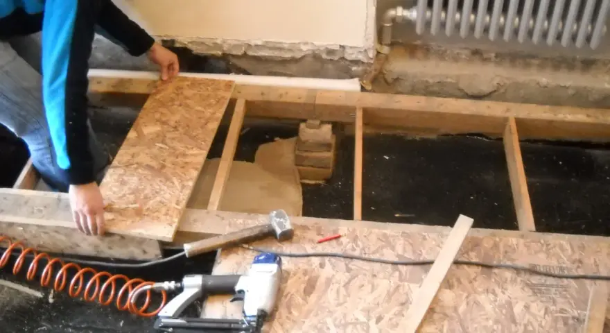 Hombre colocando tableros para nivelar suelo. Herramientas como martillo y clavadora neumática a la vista.