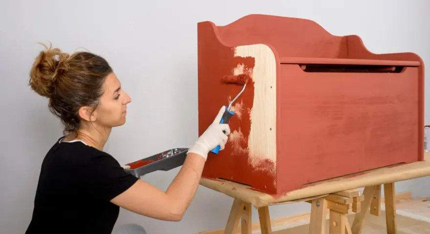 Mujer con guantes blancos pinta un mueble de madera con rodillo, cubriendo el barniz con pintura roja.