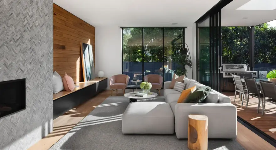 Interior de casa de madera con chimenea de mosaico, sofá modular gris, sillones rosas y acceso a terraza con barbacoa.