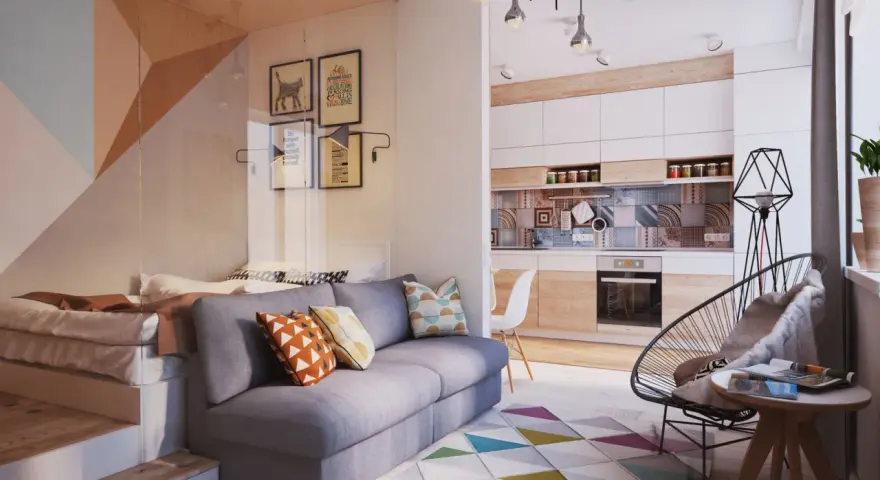 Un sofá gris con cojines coloridos. Detrás, una cama elevada y una pared con cuadros. La cocina moderna y una silla de diseño completan el espacio.