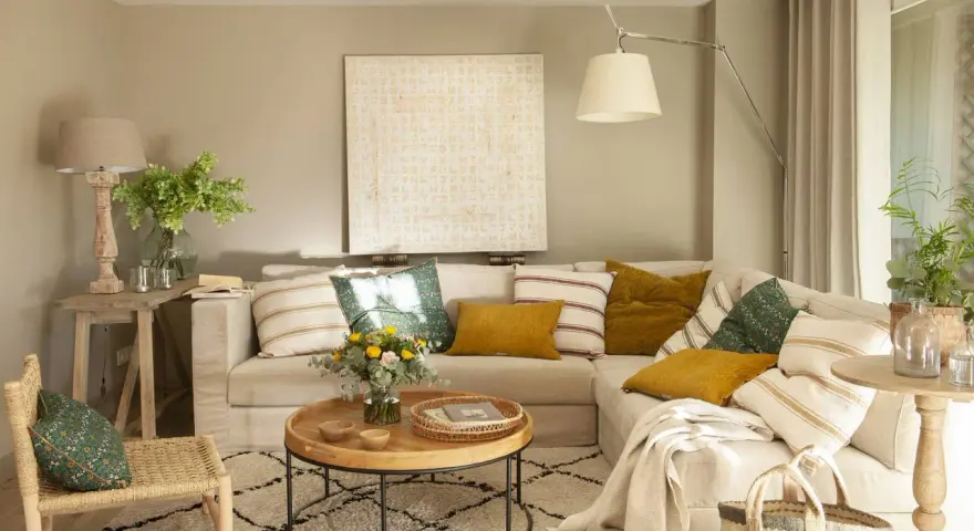 Sofá beige con cojines variados, mesa de centro redonda de madera, alfombra estampada y una silla de mimbre. El color topo cual es, se ve en las paredes y cortinas.