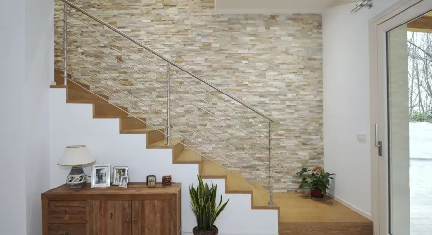 Elegante escalera con barandilla metálica y una pared de piedra natural para decorar pared subida escalera. Un mueble de madera y plantas completan la escena.