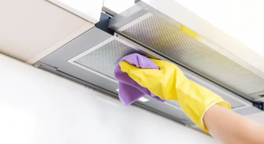 Mano con guante amarillo y paño morado limpiando el interior de la campana extractora, eliminando grasa y suciedad para un hogar reluciente.