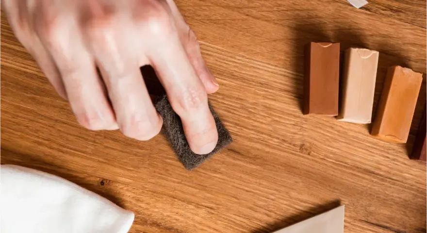 Mano lijando un parquet rayado con una esponja. Cerca, barras de cera para madera y un paño blanco.