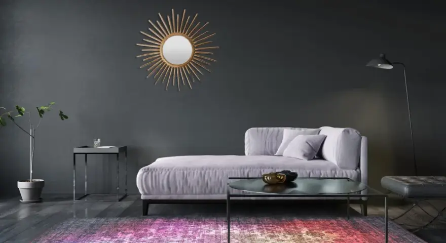 Sofá lila y alfombra vibrante sobre suelos oscuros. Un espejo sol dorado adorna la pared gris.