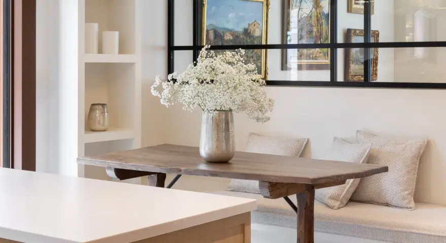 Un jarrón con flores blancas decora la mesa de madera rústica. El espacio alrededor de la mesa comedor es acogedor, con un banco tapizado y cojines.