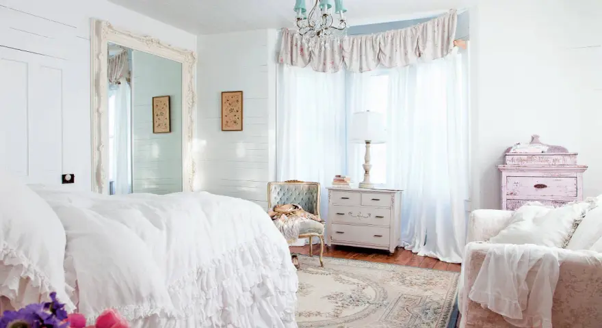 Dormitorio con cama blanca con volantes, espejo grande, cómoda antigua y un sillón. Estilo shabby chic.