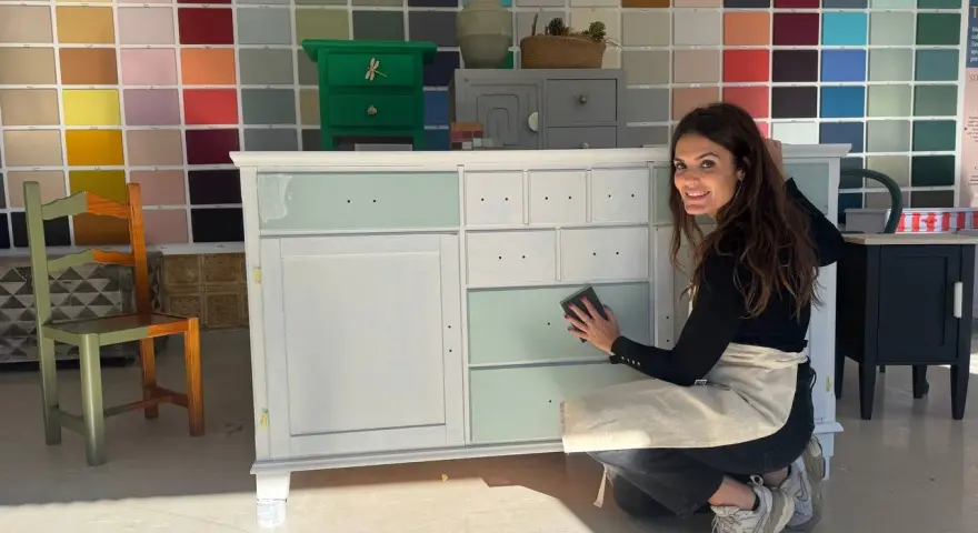 Mujer sonríe mientras lija un mueble para pintar. Detrás, una pared con muestras de colores.