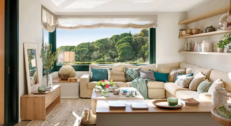 Acogedora decoracion salon comedor con sofá seccional beige, mesa de centro de madera y gran ventanal con vistas al bosque.