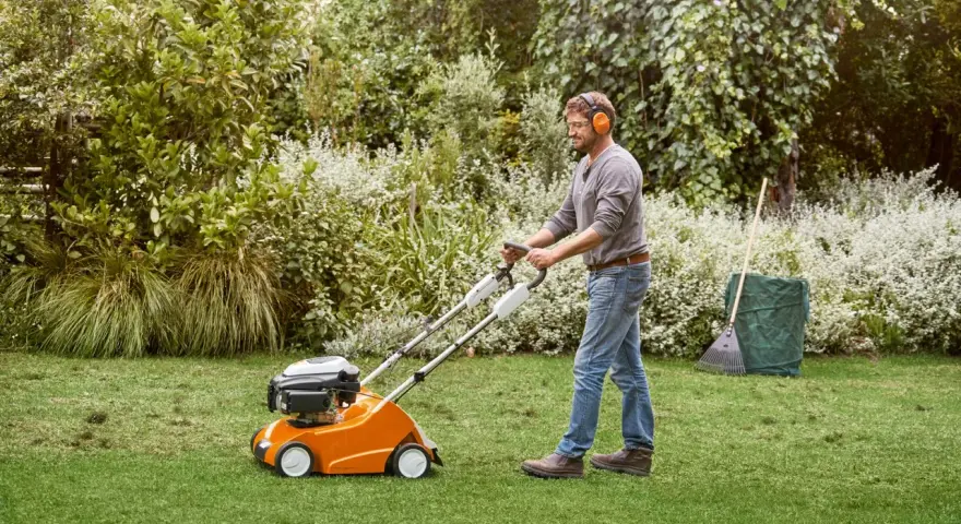 Hombre con auriculares usa un escarificador naranja para airear el césped en un jardín verde.