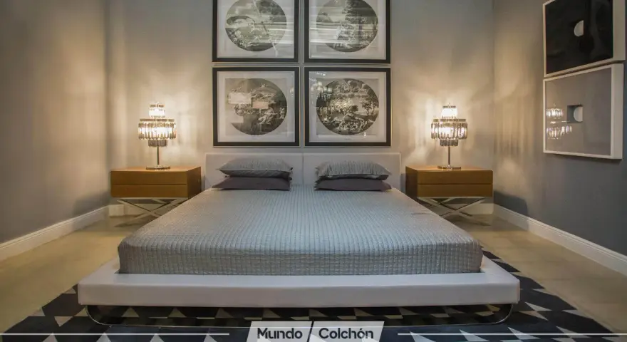 Cama moderna con cabecero tapizado, mesitas de noche de madera y lámparas de cristal. Cuadros decorativos en la pared y alfombra geométrica. Un espacio que invita a la armonía, ideal para aplicar principios de feng shui cama.