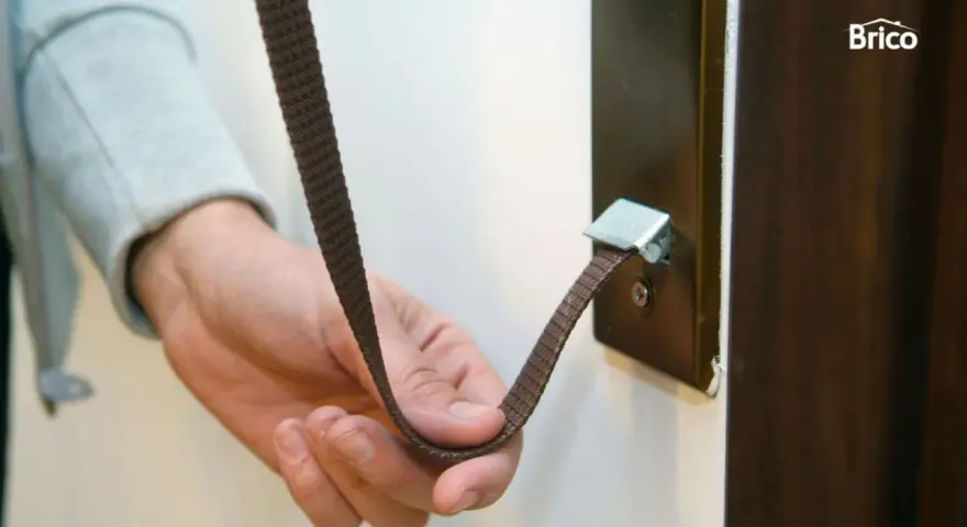 Mano sujetando una cinta marrón que se ha roto la cuerda de la persiana, enganchada a un soporte metálico.