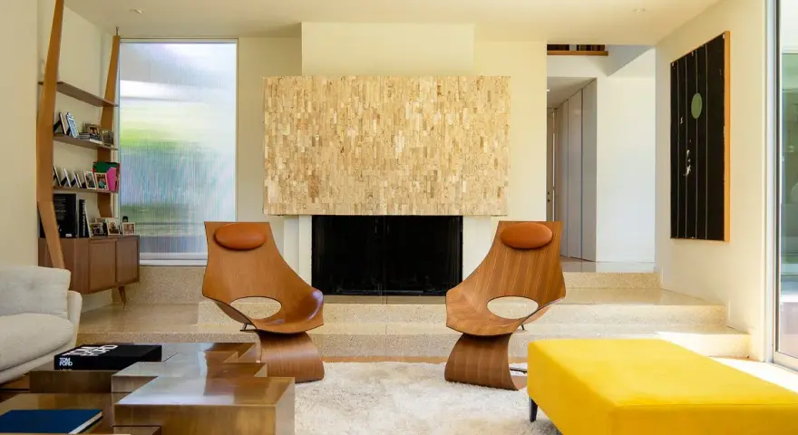 Dos sillas de madera con cojines de cuero marrón, un sofá amarillo y una chimenea de piedra, todo con un inconfundible estilo mid century.