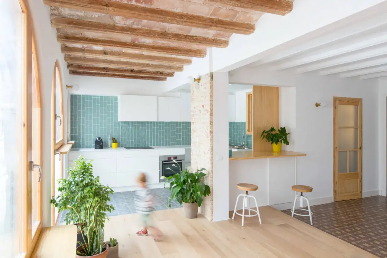 Moderna cocina tipo americana con azulejos turquesa, encimeras blancas y vigas de madera expuestas. Un espacio luminoso y abierto.