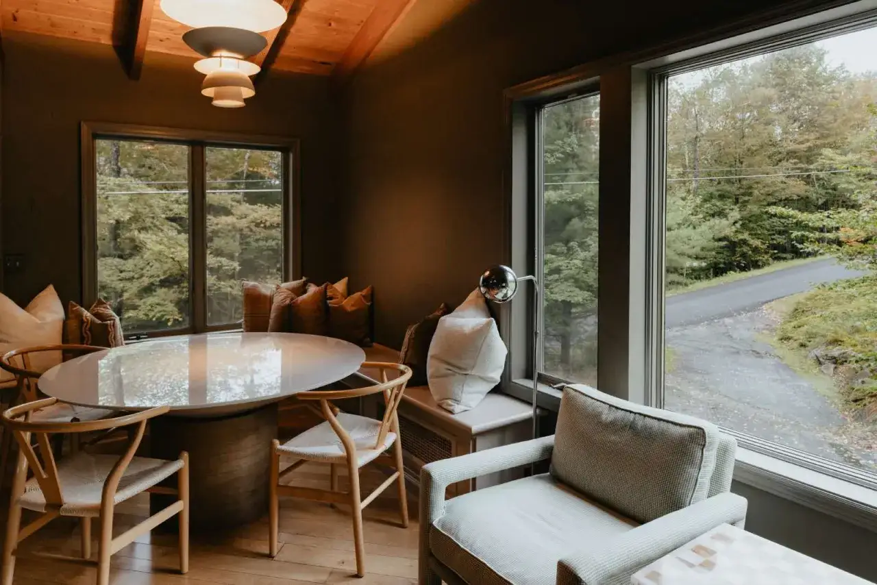 Un salón acogedor con mesa redonda y sillas de madera. La luz natural entra por grandes ventanales, ideal para saber como iluminar un salon.