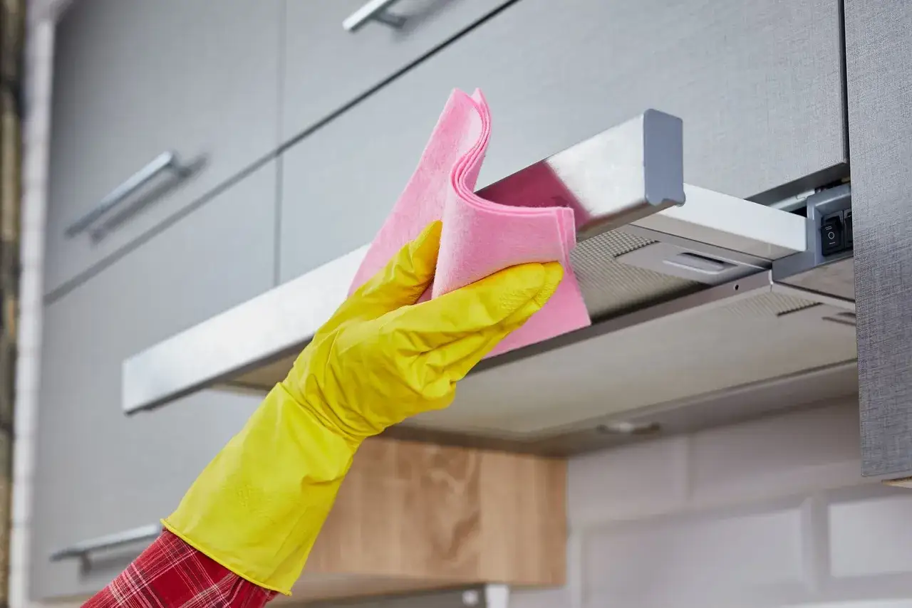Mano con guante amarillo limpiando campana de acero inoxidable con paño rosa para eliminar huellas.