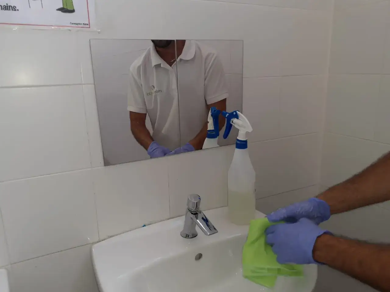 Man con guantes azules limpia el lavabo con un pa&ntilde;o verde. Botellas rociadoras y azulejos blancos en el ba&ntilde;o. Aprende como limpiar azulejos ba&ntilde;o.