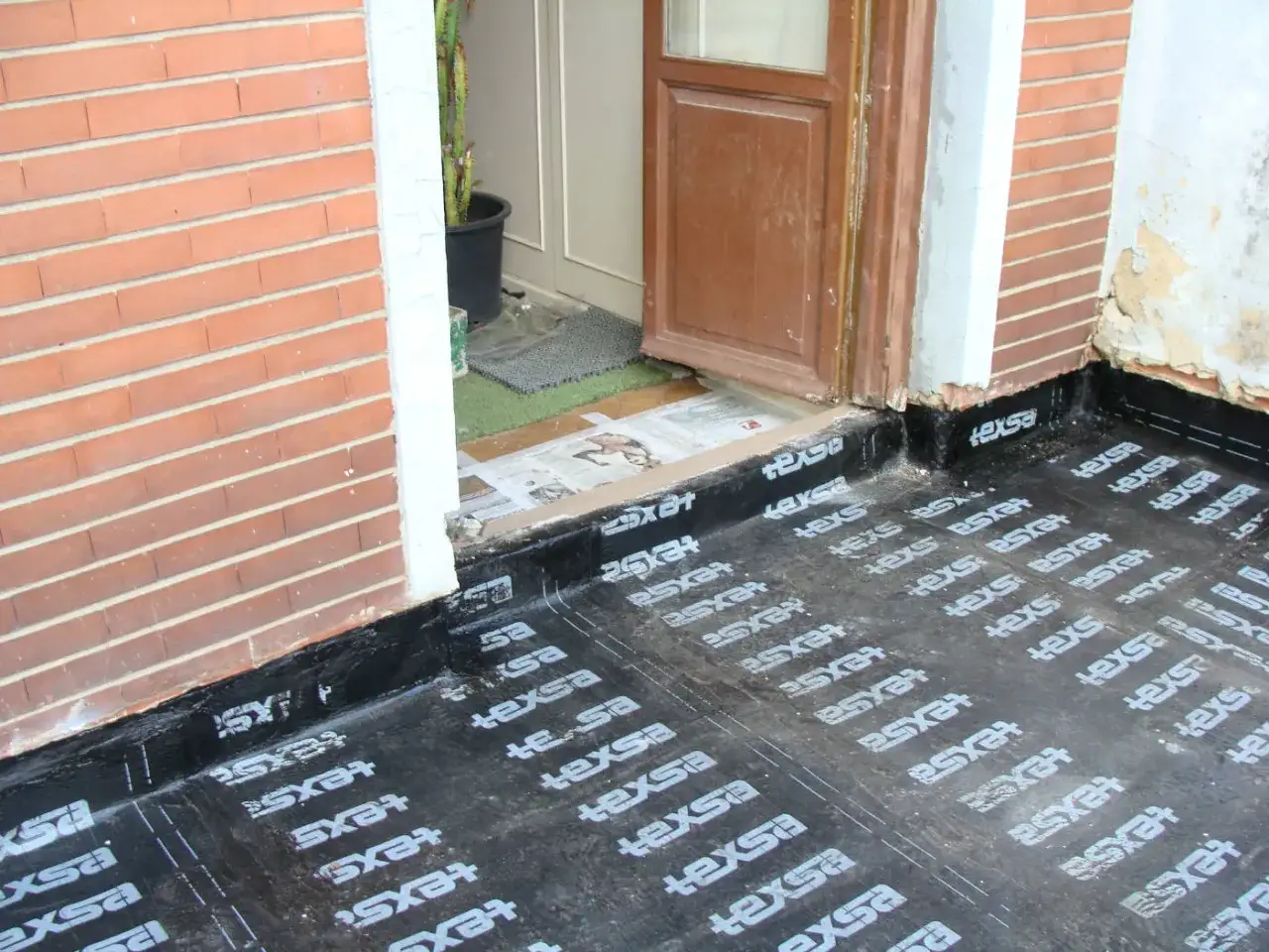 Instalaci&oacute;n de tela asf&aacute;ltica, un material impermeable, junto a una puerta y pared de ladrillo.