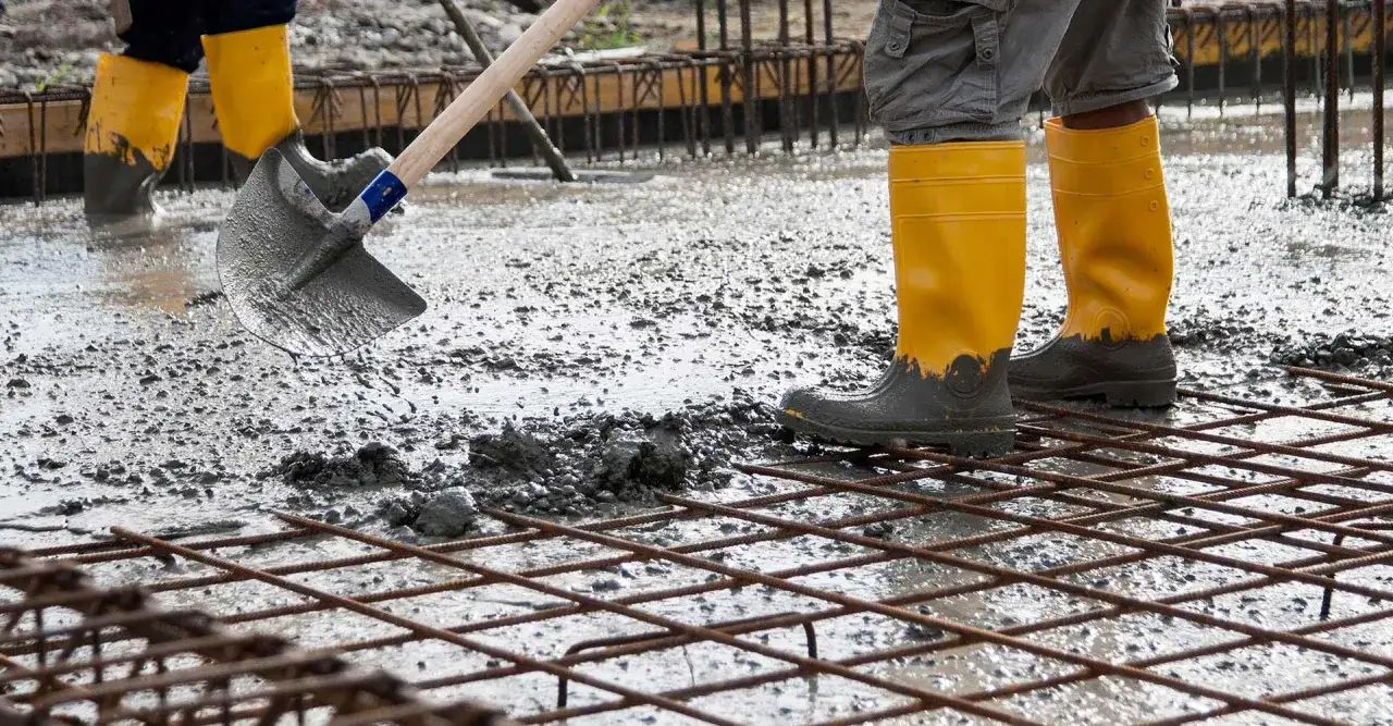 Obreros con botas amarillas trabajan en una obra, extendiendo cemento sobre una malla de ferralla.