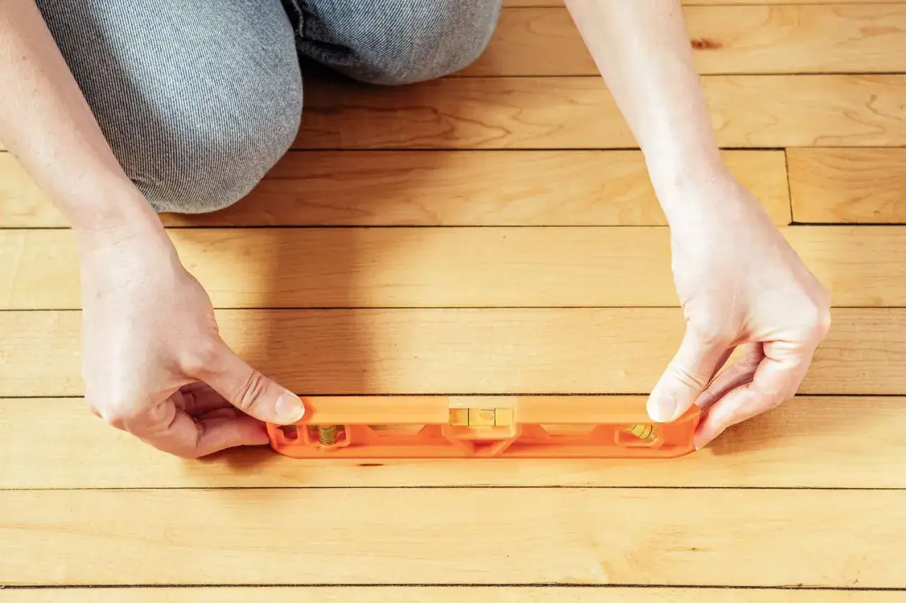 Manos sostienen nivelador naranja sobre suelo de madera antiguo para comprobar si está plano.