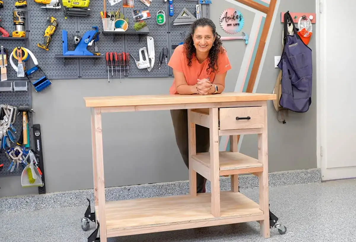 Mujer sonriente junto a un banco de trabajo de madera con caj&oacute;n y estantes. Detr&aacute;s, una pared con herramientas y sus nombres.