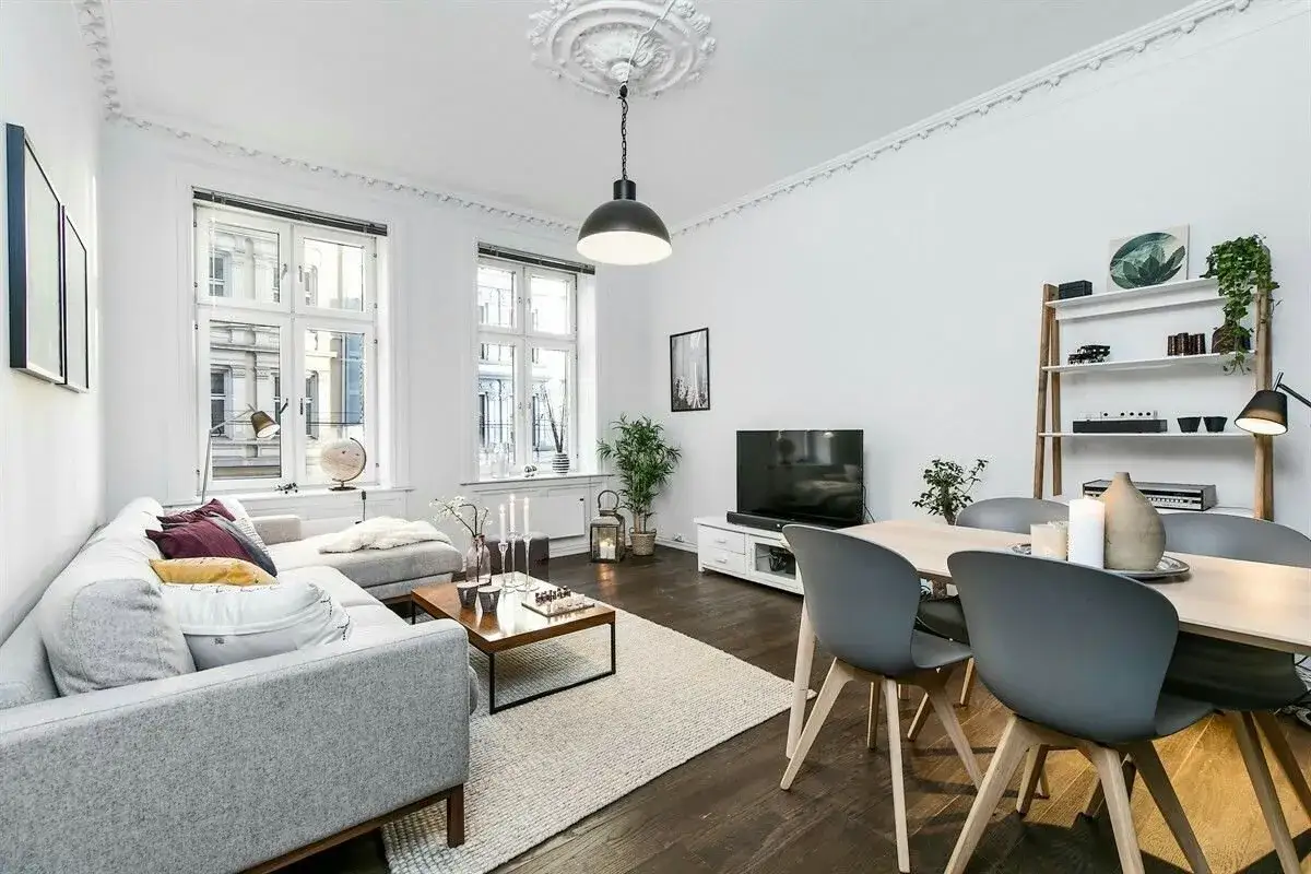 Elegante sal&oacute;n con sof&aacute;s grises, mesa de centro y suelos oscuros que contrastan con las paredes blancas y la luz natural.