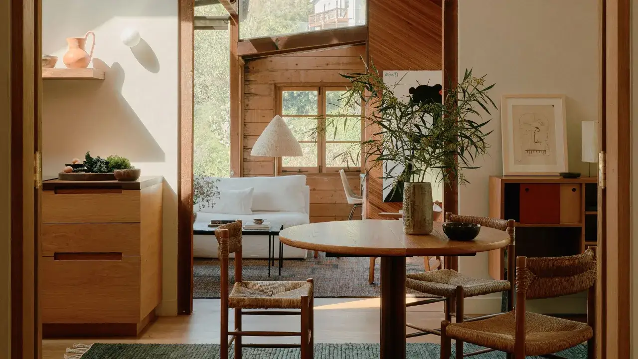 Interior casa de madera dentro, con mesa redonda, sillas de mimbre, sofá blanco y mucha luz natural.