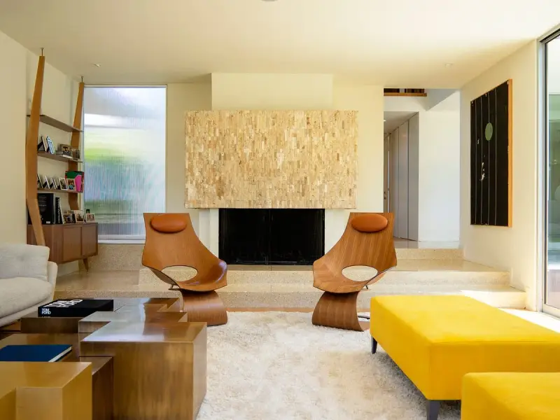 Dos sillas de madera con cojines de cuero marrón, un sofá amarillo y una chimenea de piedra, todo con un inconfundible estilo mid century.